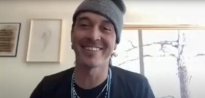A person wearing a gray beanie and black shirt is smiling at the camera. Two framed pictures are on the wall behind them, and a window is partially visible to the right.