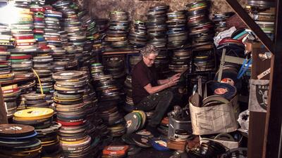 A man sits among stacks of colorful film canisters in a cluttered, dimly lit room.