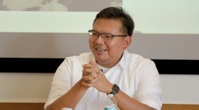 Person in a white shirt and glasses, smiling with clasped hands, a water bottle on the table.