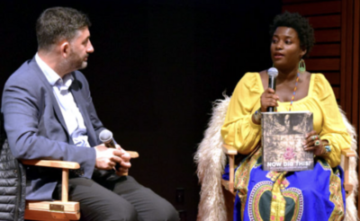 Two people seated on stage with microphones. One holds a book titled "NOW DIG THIS!".