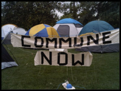 Camping tents with a banner reading "COMMUNE NOW" set up on grass.
