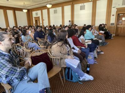 Interior of a meeting room with attendees seated in rows of wooden chairs.