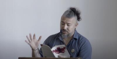 Person reading from a book at a lectern with a headset microphone.