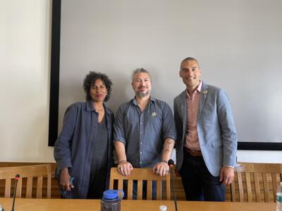 Three individuals standing side by side, smiling, with wooden chairs and a table with a blue water bottle in front of them.