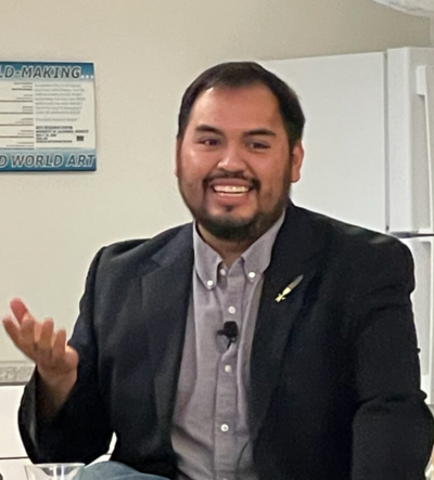 Man in a dark blazer and light gray shirt, smiling and gesturing, with a white refrigerator in the background.