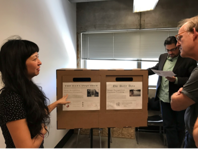 Three individuals discuss articles displayed on a cardboard stand with two sections in an office setting.