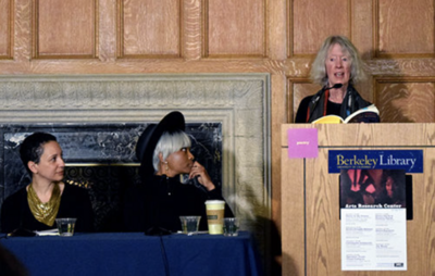  Three individuals at a poetry reading event in Berkeley Library; two seated at a table and one standing at a podium.